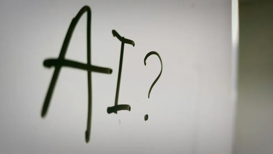 The letters AI and a question mark written in black marker on a whiteboard